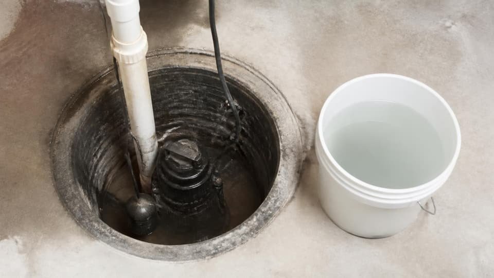A sump pump with its float switch and discharge pipe in a basement pit, with a bucket of water ready for testing